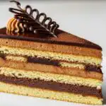 Opera Cake with thin almond sponge layers, coffee buttercream, chocolate ganache, and glossy chocolate glaze, shown in a close-up slice on a white plate with soft natural lighting.